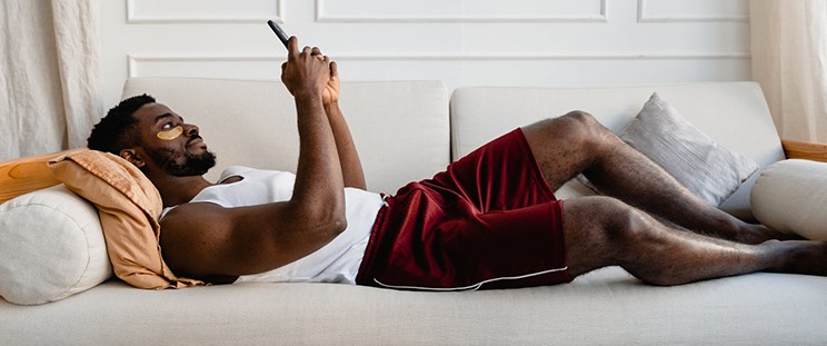 A man with an under eye patch uses his phone while lying down on the couch
