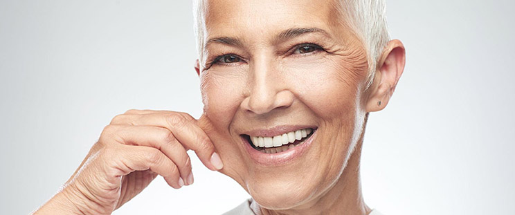 A smiling older woman pinches her cheek to show the laxity of her skin