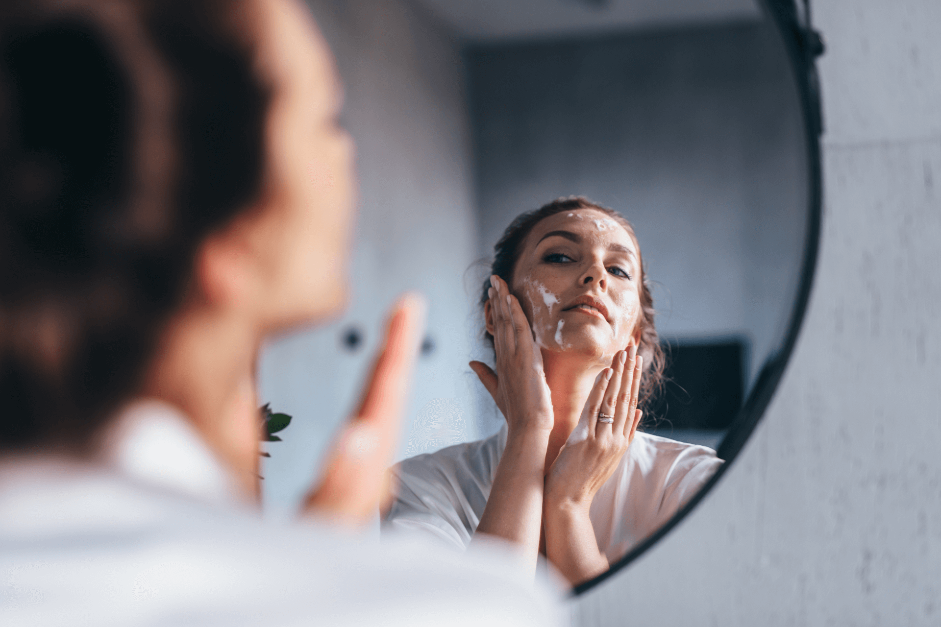 a woman applies cleanser on her face while looking in a mirror
