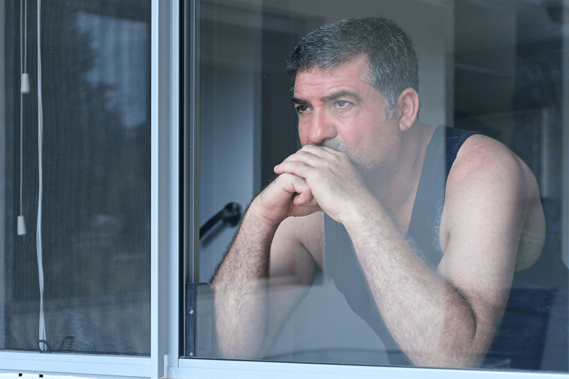 A middle-aged man in a tank top looking out a window 