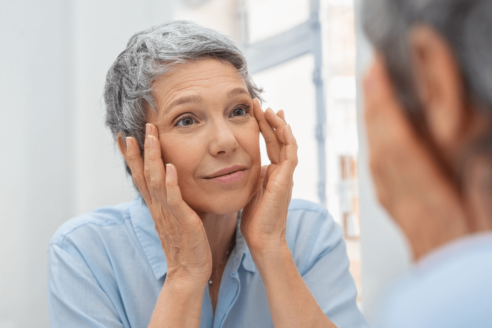 A woman with short, gray hair and forehead wrinkles checking her face in the mirror 